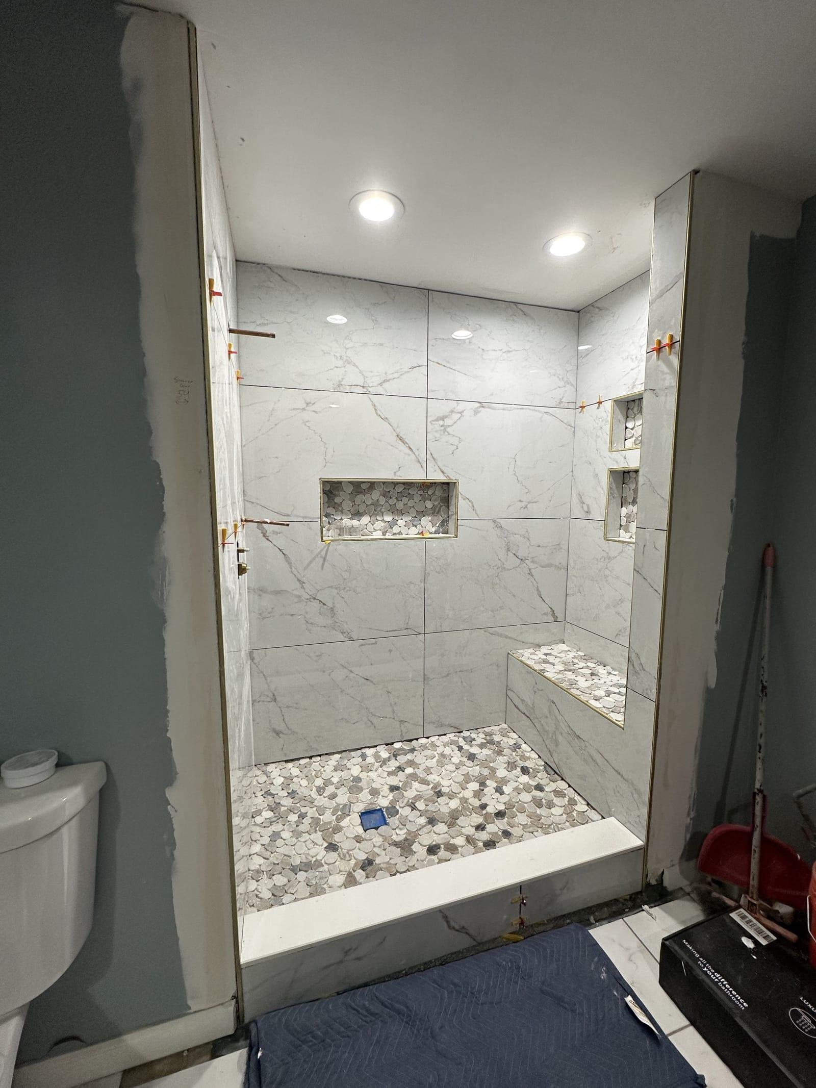 Large-format marble-look tile set with pebble floor, bench, and niches — spacers still in before grout